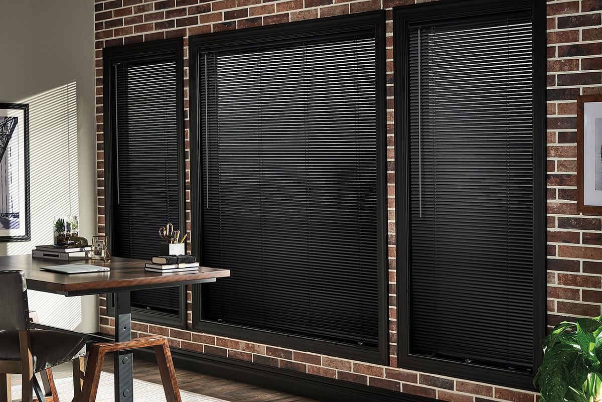 Home office with a wooden desk and black chair in front of three large windows covered by black horizontal blinds, set against a red brick wall.