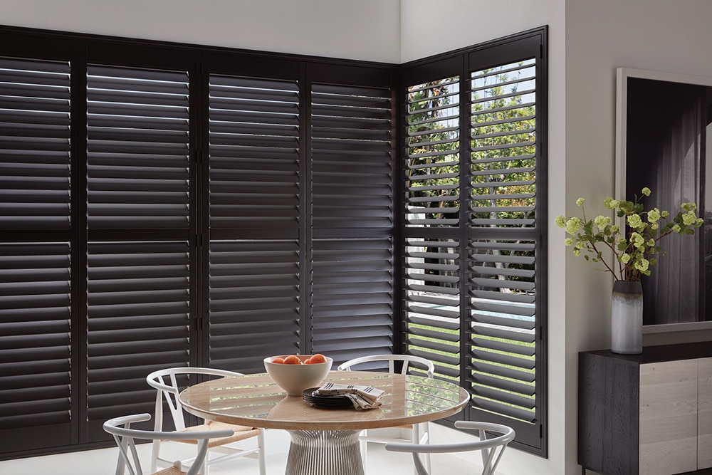 Modern dining area with round wooden table, white chairs, and large black plantation shutters partially open to reveal an outdoor view.