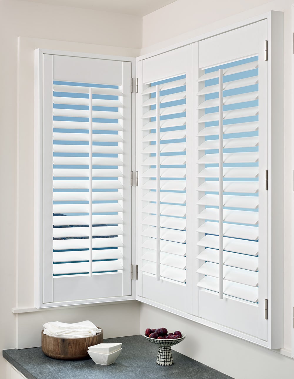 Three panels of white faux wood shutters filtering in light into a kitchen.
